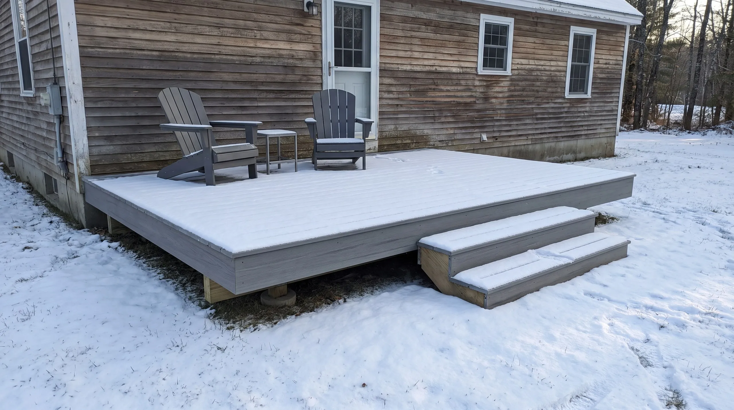 Deck contractor Litchfield Maine: low ground-level gray composite deck with two steps and Adirondack chairs, dusted with snow