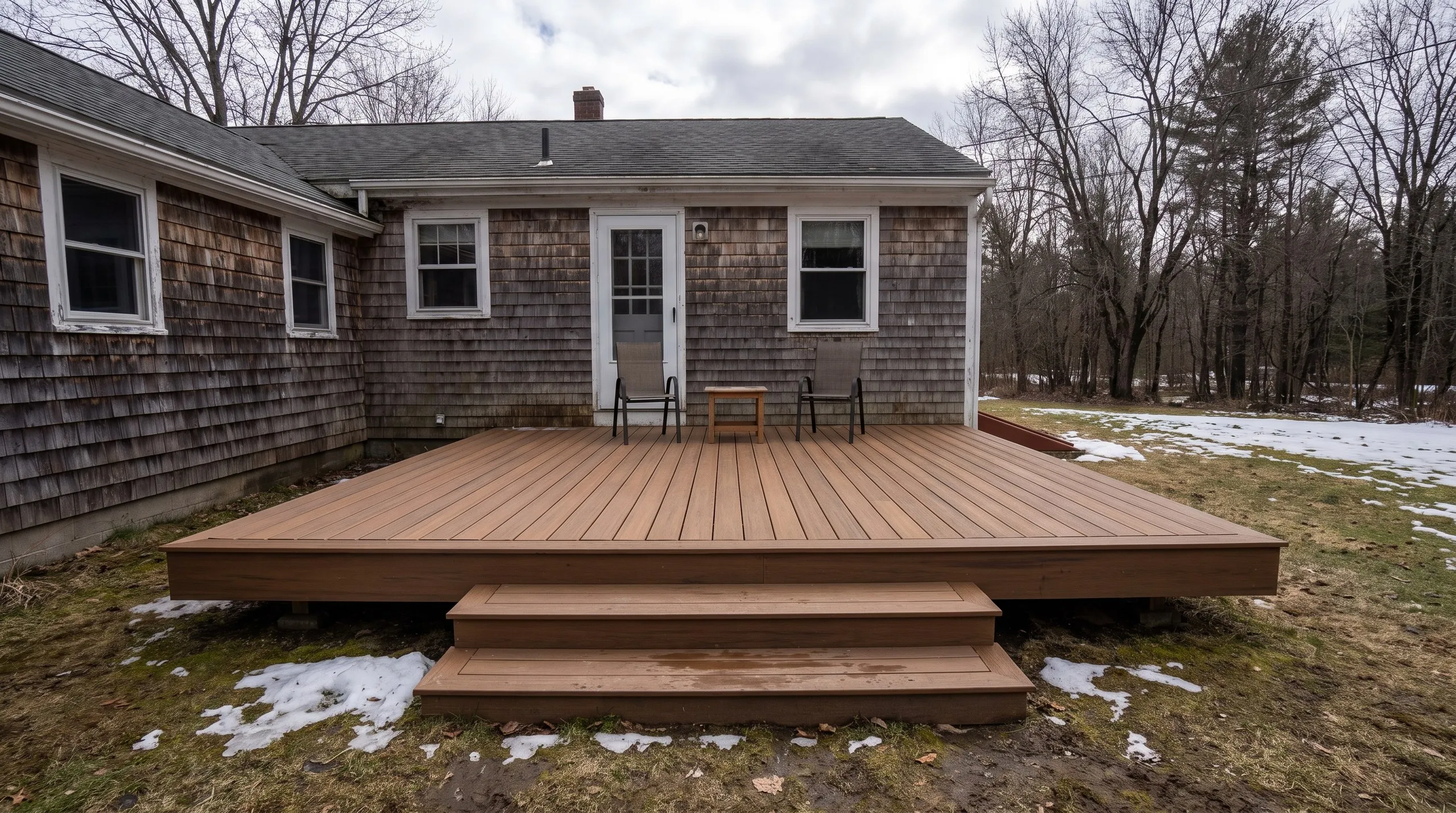 Deck contractor in Monmouth, ME: low ground-level brown composite deck with two wide steps and chairs by a shingled house