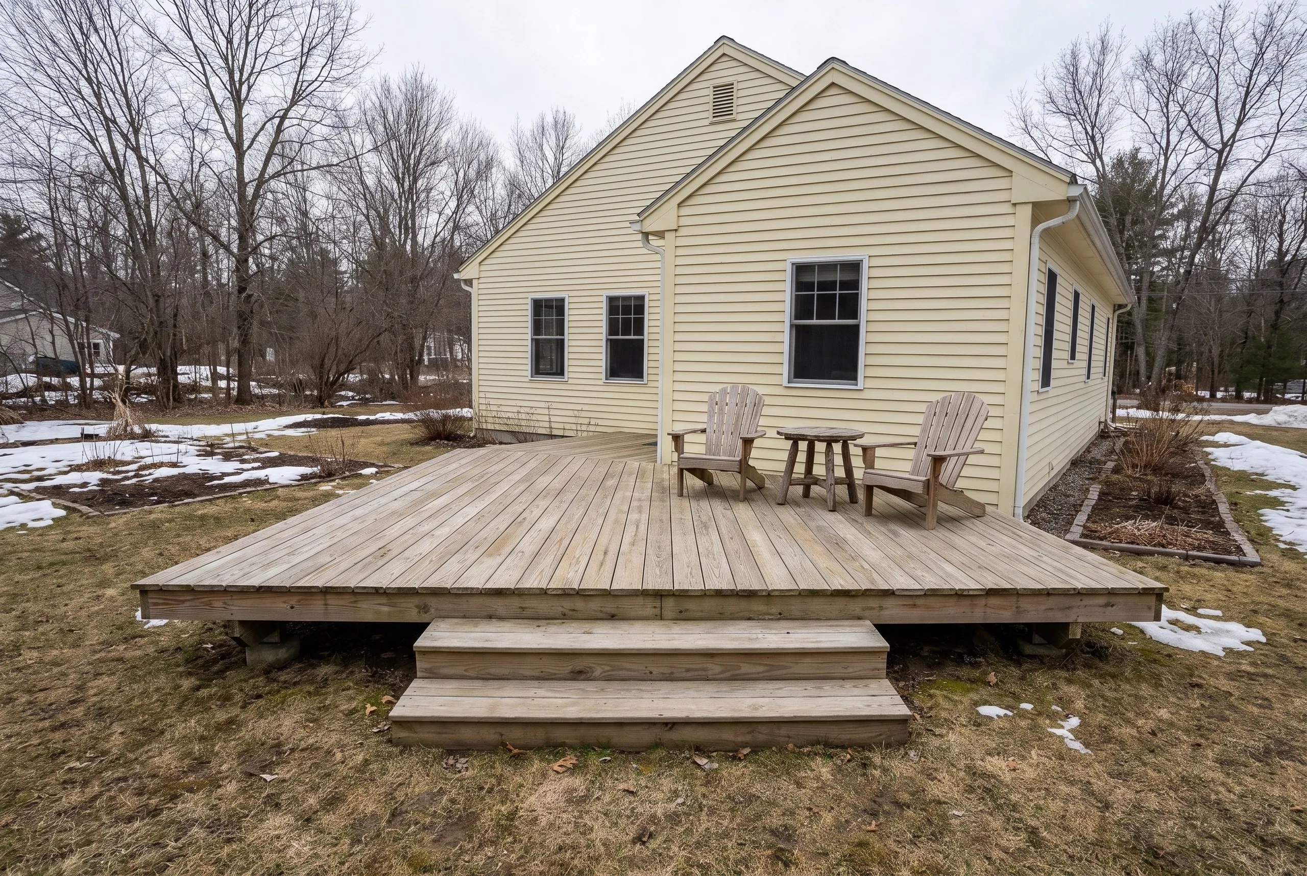 Low ground-level weathered wood deck with two wide steps and Adirondack chairs behind a pale yellow house in Monmouth, Maine