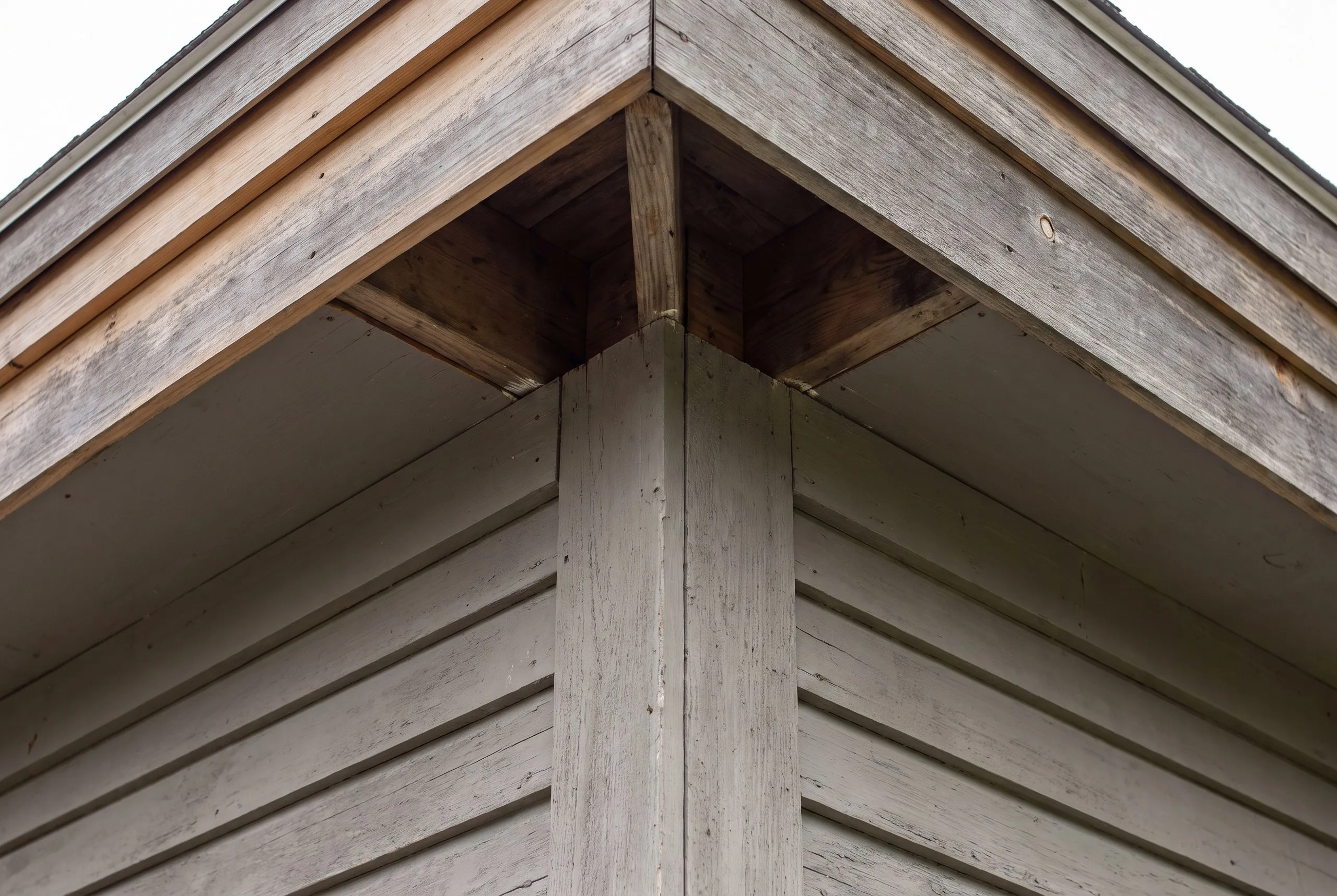Siding contractor Hallowell Maine: low view up a weathered gray corner board to the soffit and fascia, one soffit corner left open